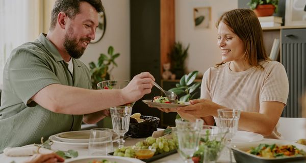 Ein Mann häuft einer Frau Salat auf den Teller, beide wirken glücklich. Auf dem Tisch steht pflanzenbetontes Essen: Salat, Trauben und ein Auflauf.