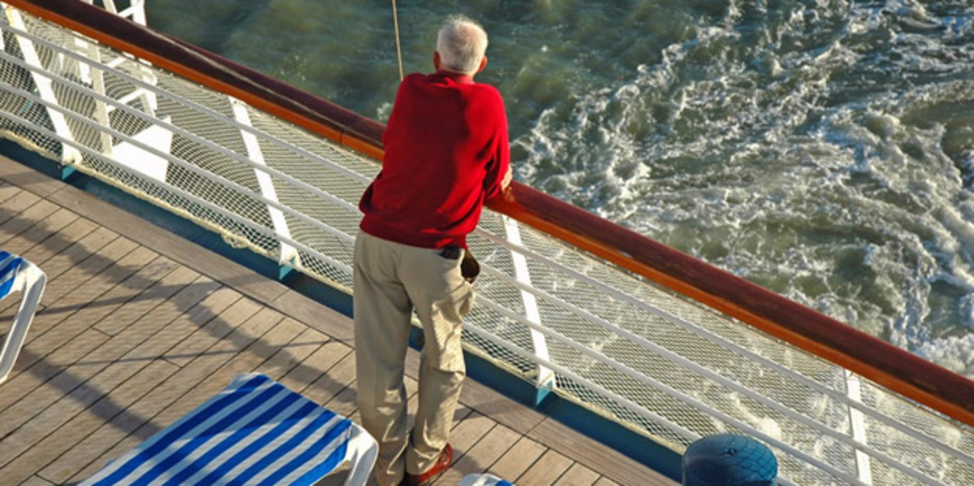 Rückenansicht eines älteren Mannes im roten Pulli und heller Hose an der Reeling eines Kreuzfahrtschiffes lehnend und aufs Meer schauend