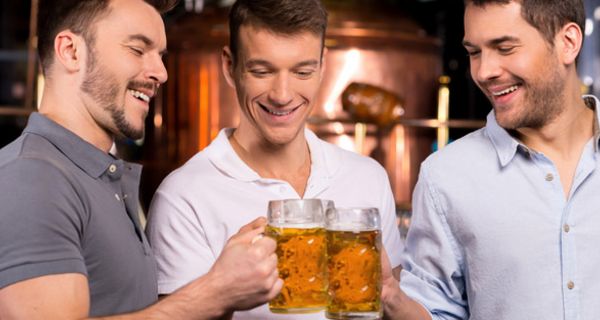 Drei Männer in den 20ern stoßen in einer Kneipe lachend mit Bierhumpen an