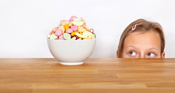 Tisch, darauf weißes Schälchen mit pastellfarbenen Süßigkeiten, Mädchen, nur bis Nasenansatz zu sehen, schaut mit verdrehten Augen hin