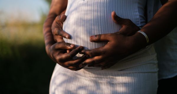 Frau mit Babybauch, wird von hinten von einem Mann umarmt, der seine Hände auf den Bauch legt.