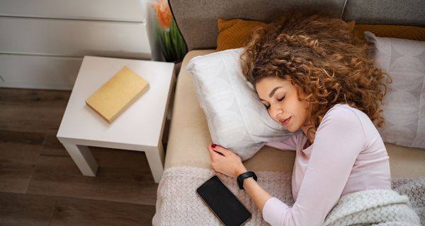 eine Frau liegt schlafend im Bett, trägt eine Uhr und neben ihr liegt ein Handy.