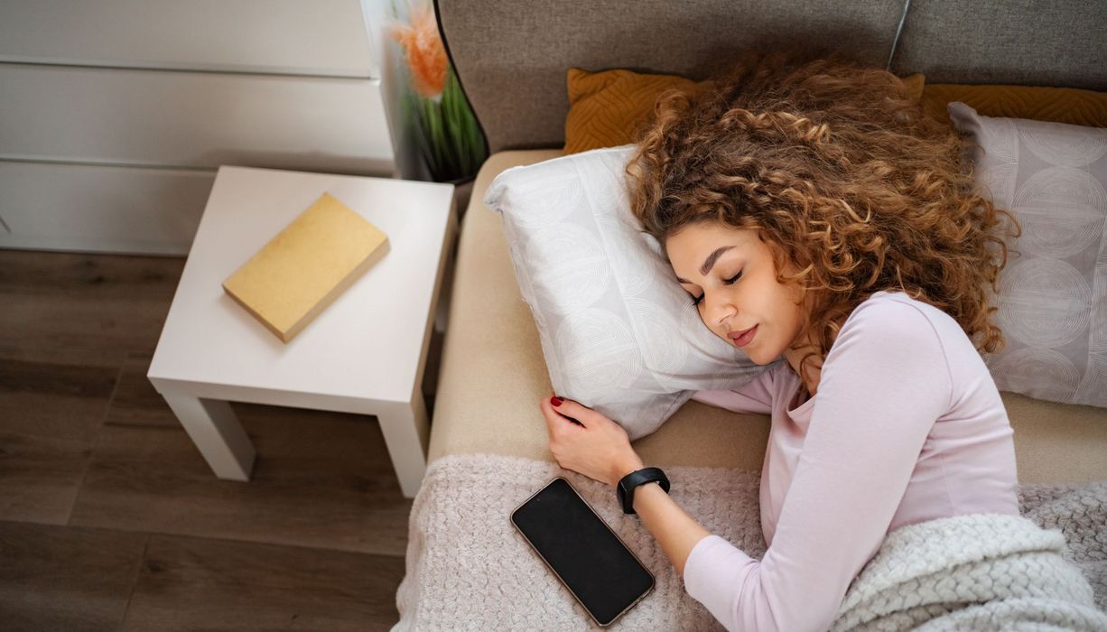 eine Frau liegt schlafend im Bett, trägt eine Uhr und neben ihr liegt ein Handy.
