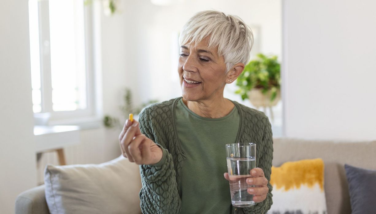 Eine ältere Frau sitzt auf dem Sofa mit einem Glas wasser in der Hand und betrachtet prüfend eine Kapsel, die sie in der anderen Hand hält.