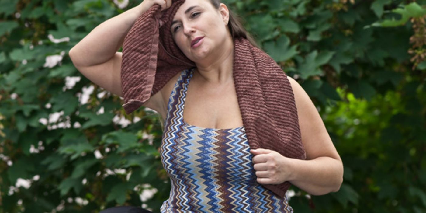 Übergewichtige jüngere Frau in sommerlicher Sportkleidung im Freien wischt sich mit Handtuch Schweiß aus dem Gesicht nach dem Sport