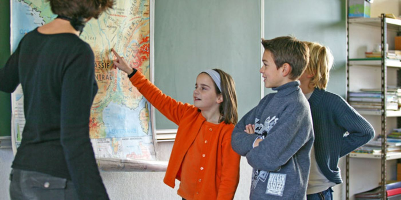 Lehrerin (Rückansicht) mit Grundschülerin und 2 Schülern an einer Landkarte. Schülerin zeigt mit dem Finger auf etwas auf der Karte