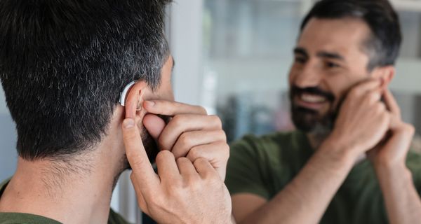 Ein Mann steht vor einem Spiegel. Er lächelt und hat eine Hand an einem Hörgerät in seinem Ohr. 