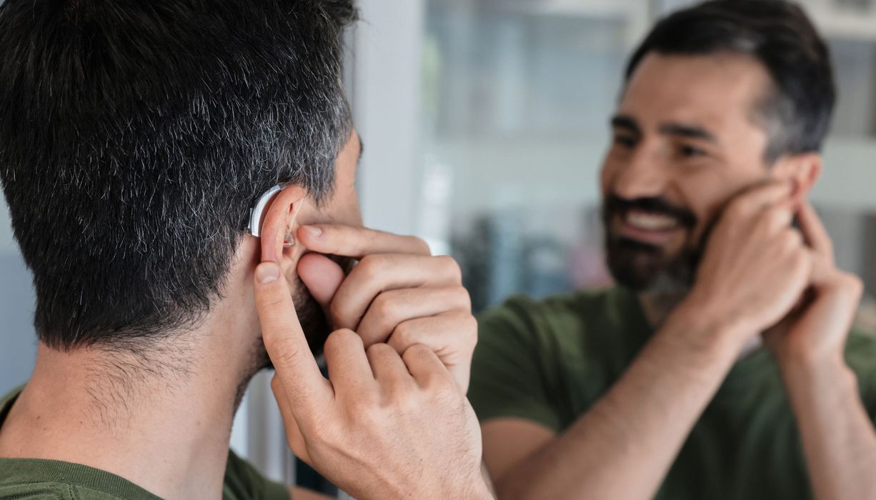 Ein Mann steht vor einem Spiegel. Er lächelt und hat eine Hand an einem Hörgerät in seinem Ohr. 