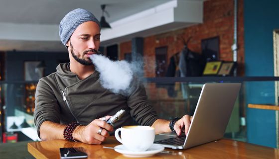 Junger Mann am Laptop nutzt eine E-Zigarette.