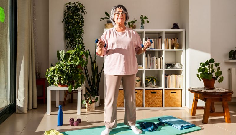 Ältere Frau mit Brille steht auf einer Yogamatte, zwei kleine Hanteln in den Händen, entschlossener Gesichtsausdruck.