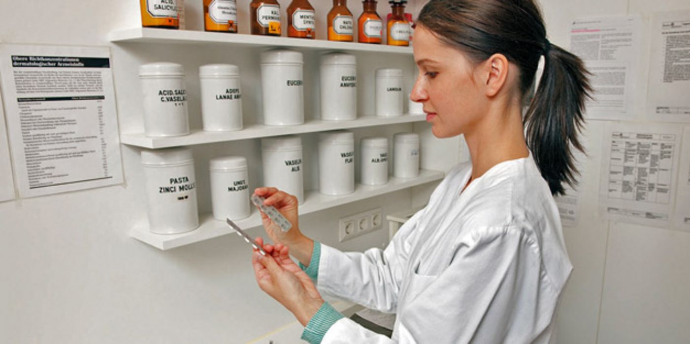 Junge Apothekerin schaut prüfend auf Blisterpackungen, auf einem Regal alte Arzneigefäße aus Porzellan und Glas