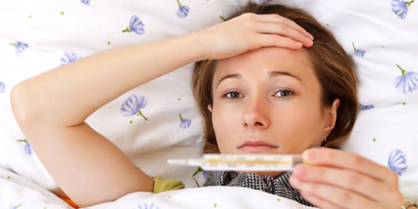 Junge Frau im Bett schaut auf Fieberthermometer und fasst sich an den Kopf