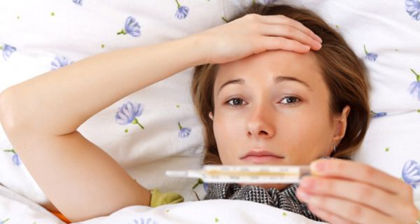 Junge Frau im Bett schaut auf Fieberthermometer und fasst sich an den Kopf