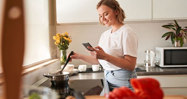 Junge Frau, kocht eine Suppe und schaut auf ihr Smartphone.