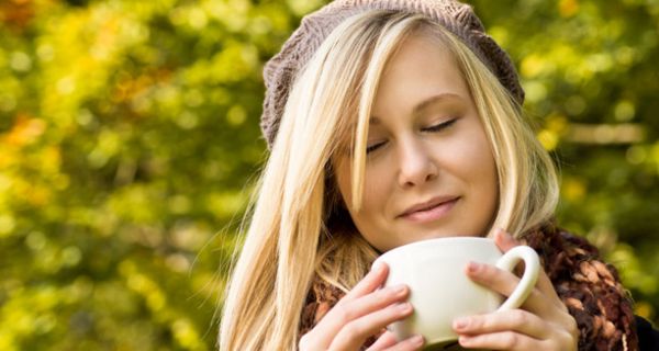 Blonde junge Frau, ca. 18, hellbraune Wollmütze, blonde halblange Haare, schaut genüsslich in eine Kaffeetasse; im Hintergrund herbstlicher Blätterwald