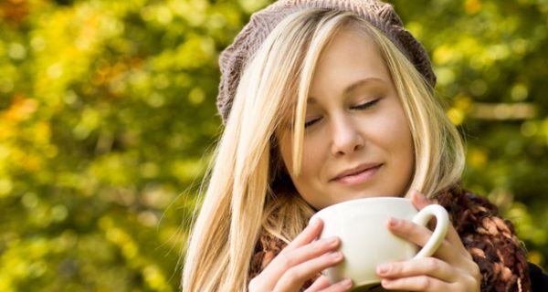 Blonde junge Frau, ca. 18, hellbraune Wollmütze, blonde halblange Haare, schaut genüsslich in eine Kaffeetasse; im Hintergrund herbstlicher Blätterwald