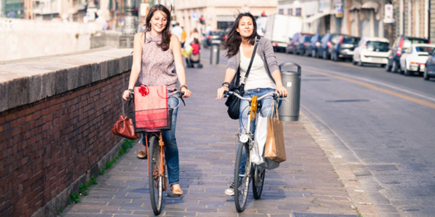 Sommerszene: 2 junge Frauen, vom Einkaufen kommend, fahren in einer Stadt auf einem gepflasterten Weg auf Rädern nebeneinander her auf die Kamera zu