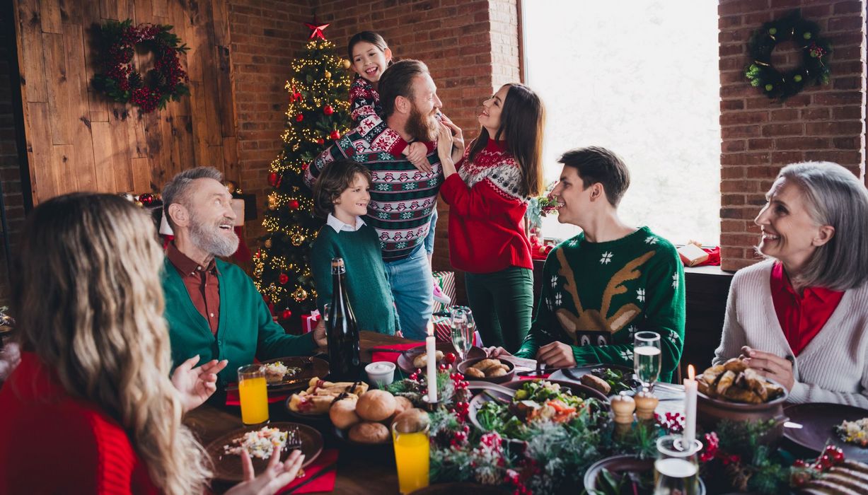 Acht Personen rund um den gedeckten Weihnachtstisch: Ein Kind sitzt auf den Schultern eines Mannes. Eine ältere Frau sitzt am Rand.