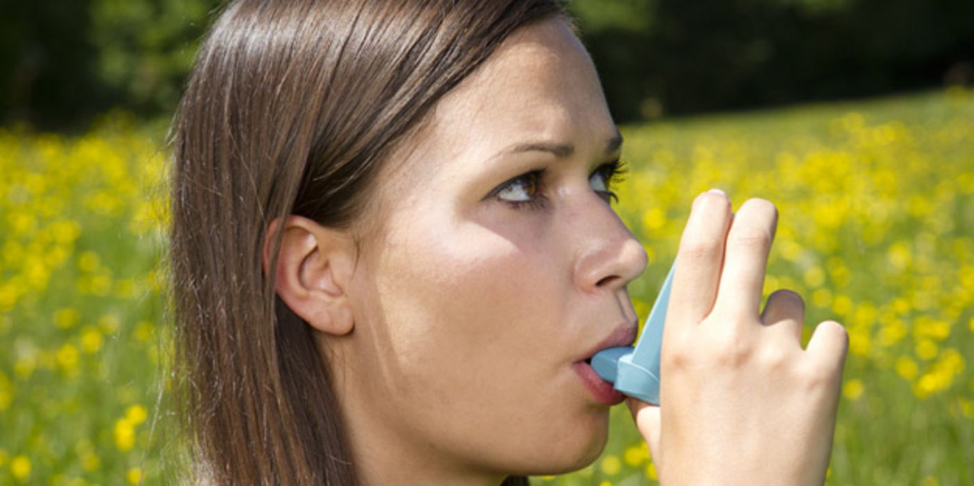 Junge Frau mit Asthmaspray im Mund auf einer Wiese mit Löwenzahn