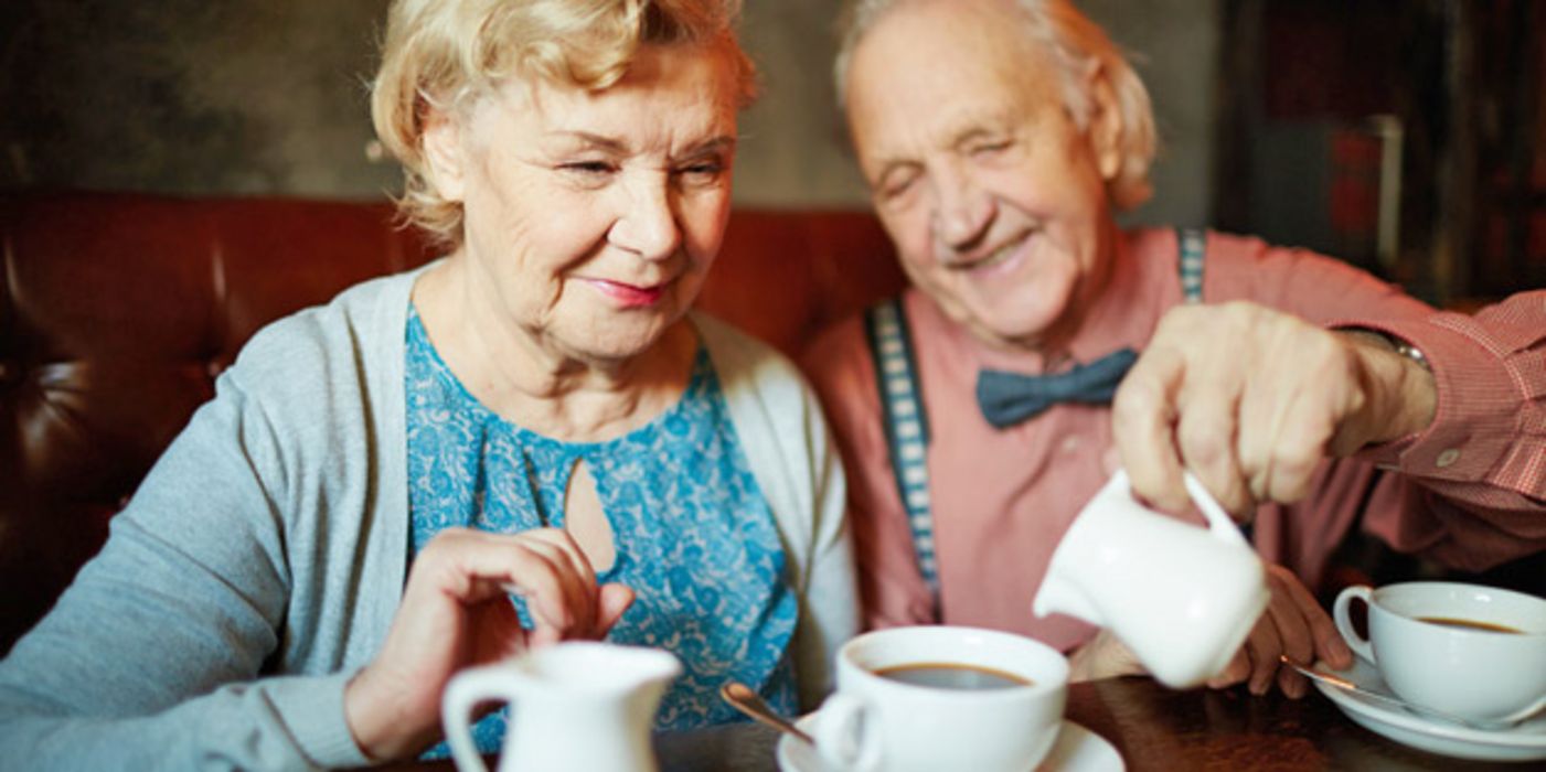 Seniorenpaar, gefüllte Kaffeetassen vor sich; Frau verschmitzt in Kamera schauend, Mann schenkt ihr Milch in den Kaffee ein