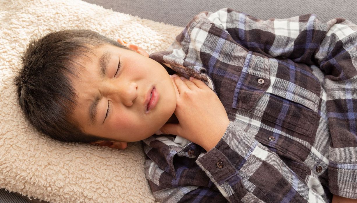 Junge, etwa acht Jahre alt, fasst sich an den Hals.