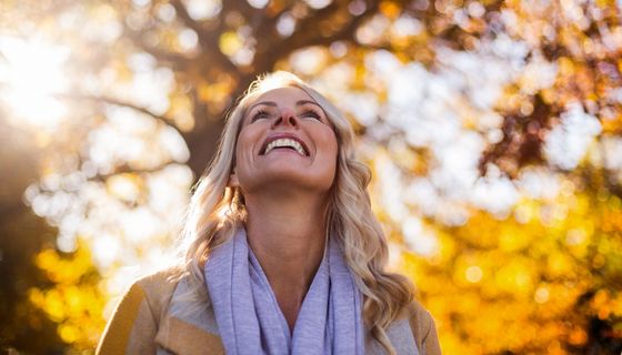 Ältere Frau, steht unter Herbstbäumen und schaut lächelnd nach oben.