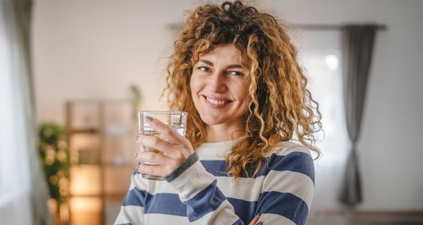 Frau mit blonden Locken hält ein Glas Wasser in der Hand.