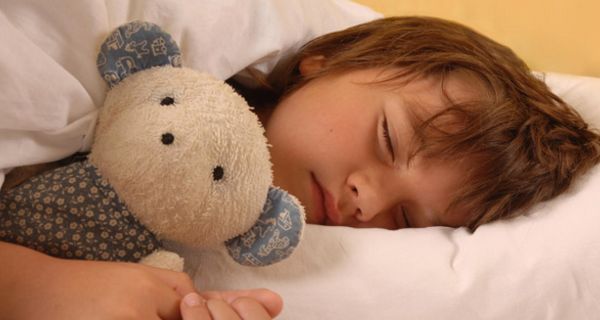 Kleines Mädchen, braune Haare, liegt auf einem weißen Kopfkissen mit einem Kuscheltier und schläft.