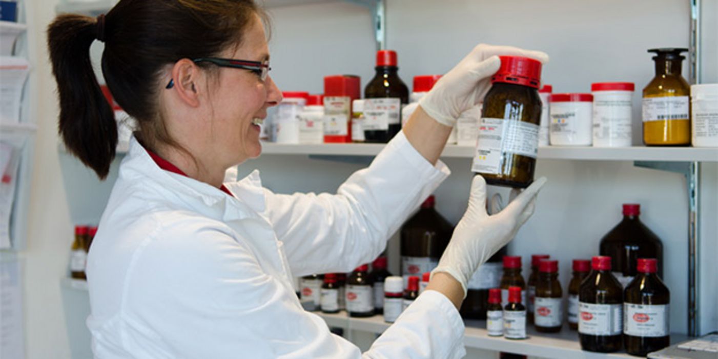 Apothekerin im Laborbereich, lachend, Schutzhandschuhe, schaut auf Etikett einer braunen Glasflasche, die sie aus Regal genommen hat