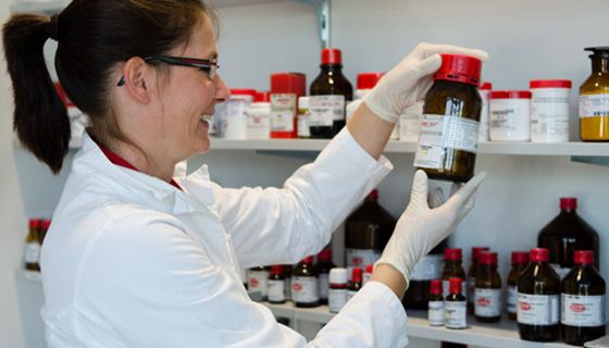 Apothekerin im Laborbereich, lachend, Schutzhandschuhe, schaut auf Etikett einer braunen Glasflasche, die sie aus Regal genommen hat