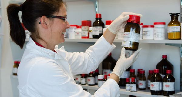 Apothekerin im Laborbereich, lachend, Schutzhandschuhe, schaut auf Etikett einer braunen Glasflasche, die sie aus Regal genommen hat