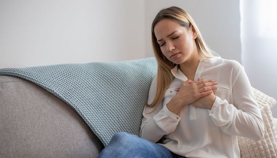 Junge Frau sitzt auf dem Sofa, beide Hände auf die Brust gedrückt, das Gesicht schmerzhaft verzogen.