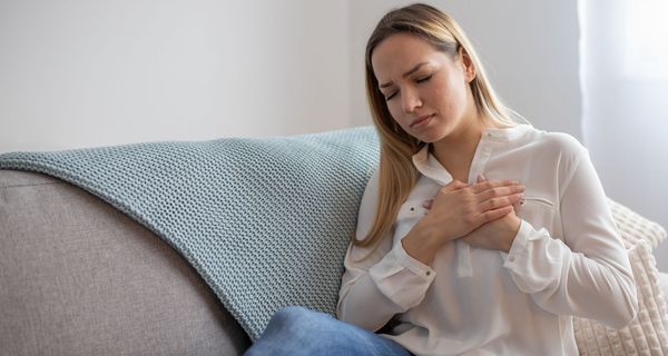 Junge Frau sitzt auf dem Sofa, beide Hände auf die Brust gedrückt, das Gesicht schmerzhaft verzogen.
