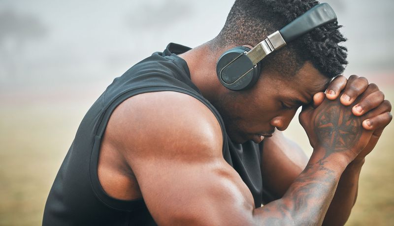 Muskulöser Mann sitzt in sich zusammengefaltet da, die Stirn auf verschränkten Händen, im Hintergrund eine neblige Landschaft.