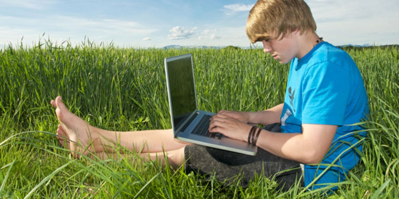 Blonder Teenagerjunge, ca. 15,  in blauem Shirt auf grüner Wiese mit einem Laptop auf den Beinen
