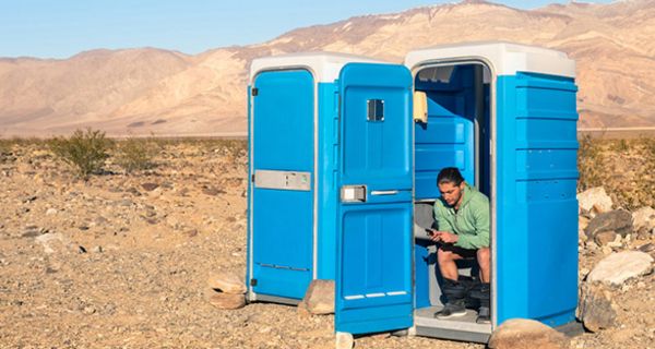 Kalifornische Wüste: Mann, ca. 30, in Kakihemd, runtergelassene Hose, bei offener Tür auf Toilette in blauem Pixiklo sitzend, sms schreibend