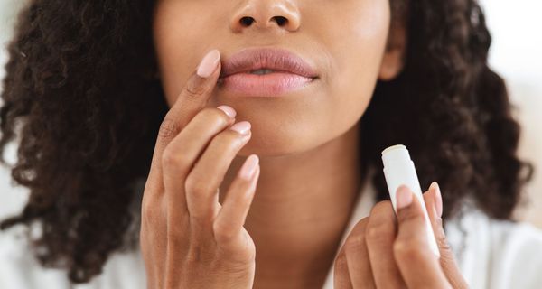 Dunkelhäutige Frau, hält Lippenpflegestift in der einen Hand und trägt diesen mit der anderen Hand auf.