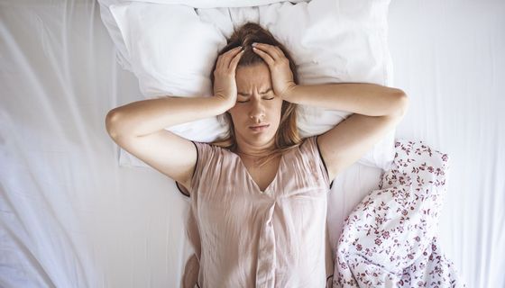 Frau, liegt im Bett und hält sich den Kopf.