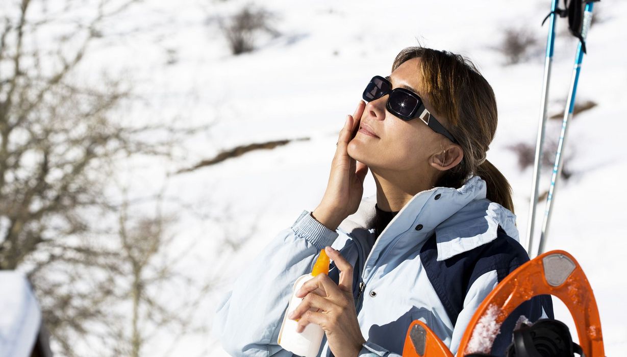 Frau in Winterkleidung und mit Schneeschuhen verteilt Sonnencreme in ihrem Gesicht.