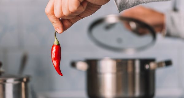 Frau, hält eine Chili in die Kamera, im Hintergrund ist ein Kochtopf.