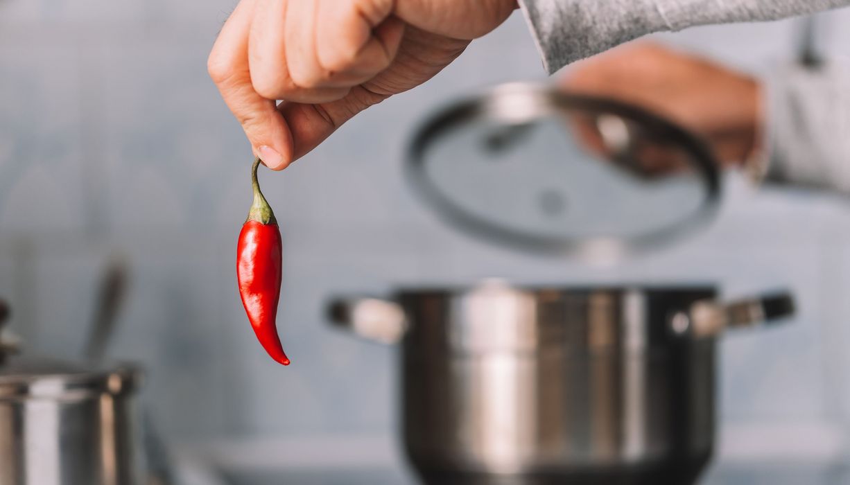 Frau, hält eine Chili in die Kamera, im Hintergrund ist ein Kochtopf.