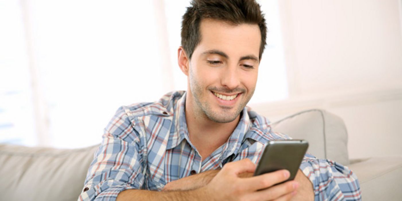 Junger Mann sitzt auf einer Couch und bedient ein Smartphone