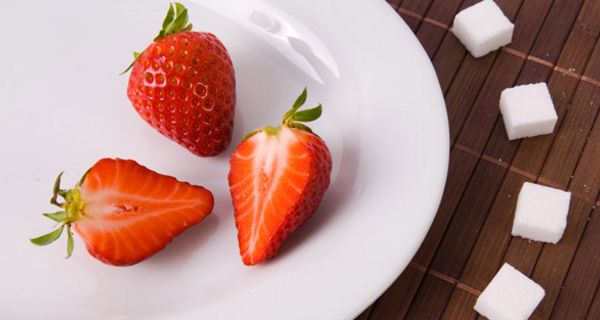 Großaufnahme, links angeschnittener weißer Teller, darauf 3 halbe Erdbeeren, 2 davon mit Innenseite nach oben, darum gelegt auf dunkler Fläche 5 Zuckerwürfel