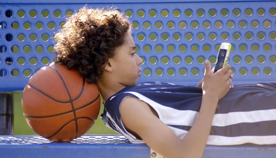 Junge, liegt mit Smartphone auf einer Bank und stützt seinen Kopf auf einen Basketball.