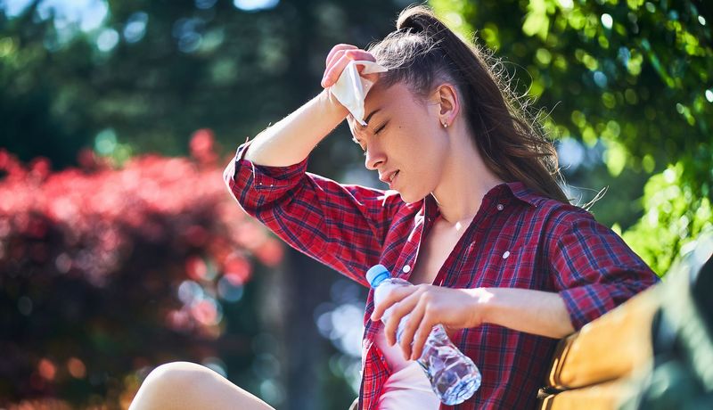Junge Frau sitzt auf einer Bank, wischt sich den Schweiß von der Stirn und hält Wasser in einer Flasche bereit.