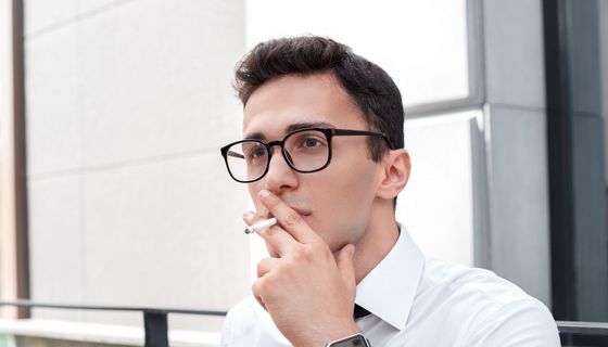 Junger Mann mit Brille im Hemd raucht eine Zigarette, an ein Geländer gelehnt.