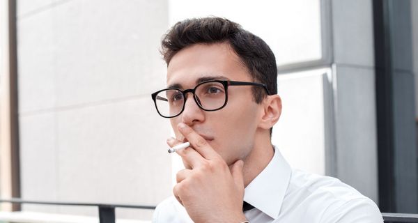 Junger Mann mit Brille im Hemd raucht eine Zigarette, an ein Geländer gelehnt.