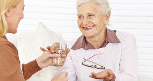 Lächelnde ältere Dame mit Tablette im linken Handteller bekommt von jüngerer Frau ein Glas Wasser gereicht