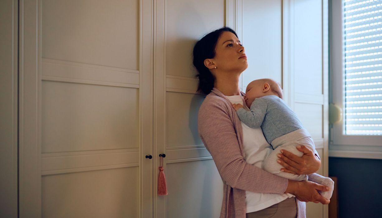 Frau, lehnt sich verzweifelt mit Baby im Arm an einen Schrank.