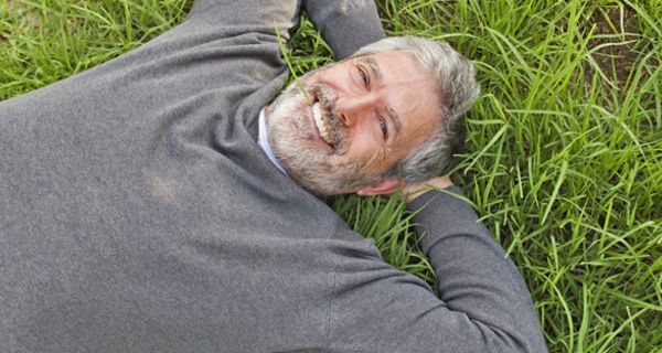 Foto von oben: Mann in den 50ern, lachend, im Gras liegend; graumelierter Bart und Haare, grauer V-Ausschnitt-Pullover, Arme hinter dem Kopf verschränkt